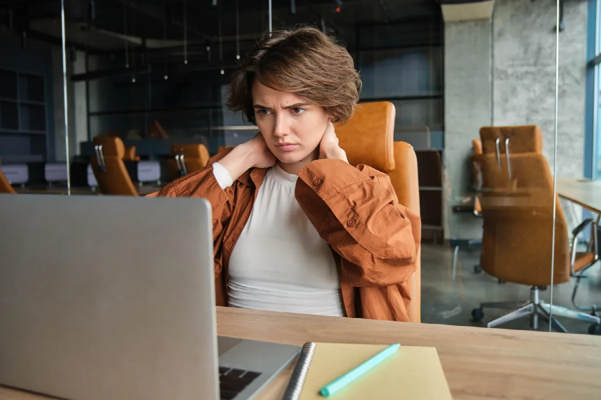 Eine junge Frau mit kurzen braunen Haaren sitzt im Büro vor ihrem Laptop und greift sich mit angespannter Miene an den Nacken.