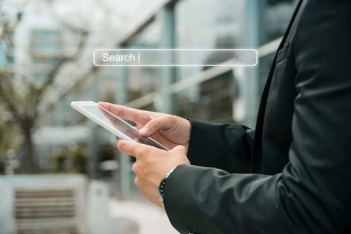 Eine Person im Anzug bedient draußen ein weißes Smartphone; darüber ist eine leuchtende Suchleiste mit dem Wort „Search |“ eingeblendet.